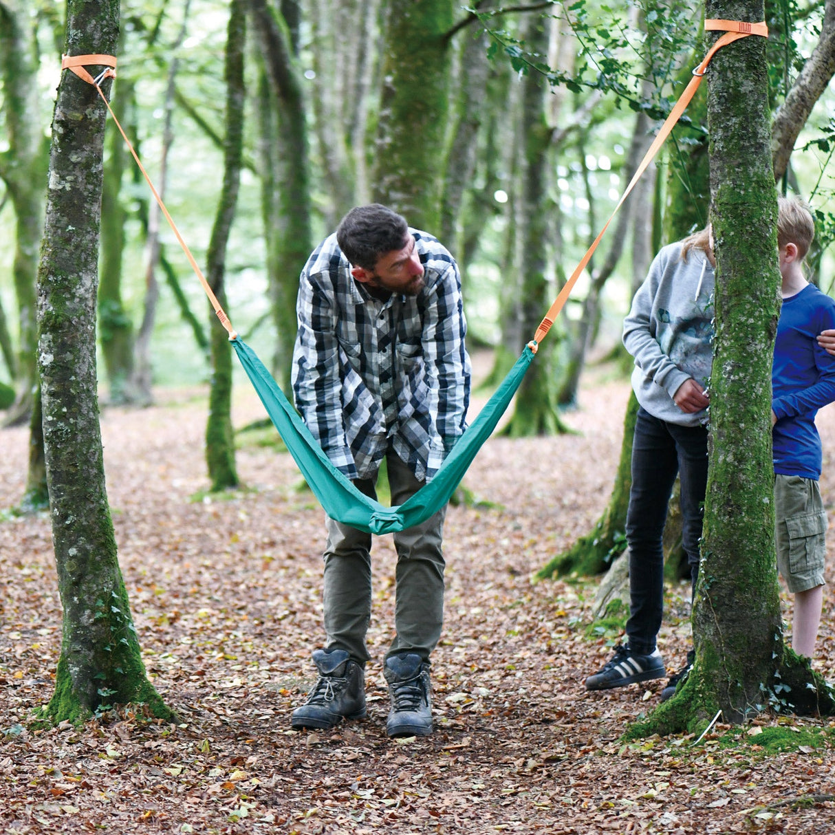 Man setting up the Pocket Swing between two trees in a forest while children watch nearby.