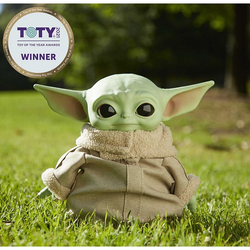Star Wars The Child plush toy sitting on grass wearing beige jacket with award badge in background.