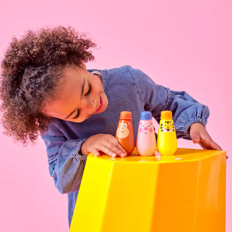 Child playing with colorful animal-shaped bubble bottles Squeezy Bubbles on a yellow stool against pink background