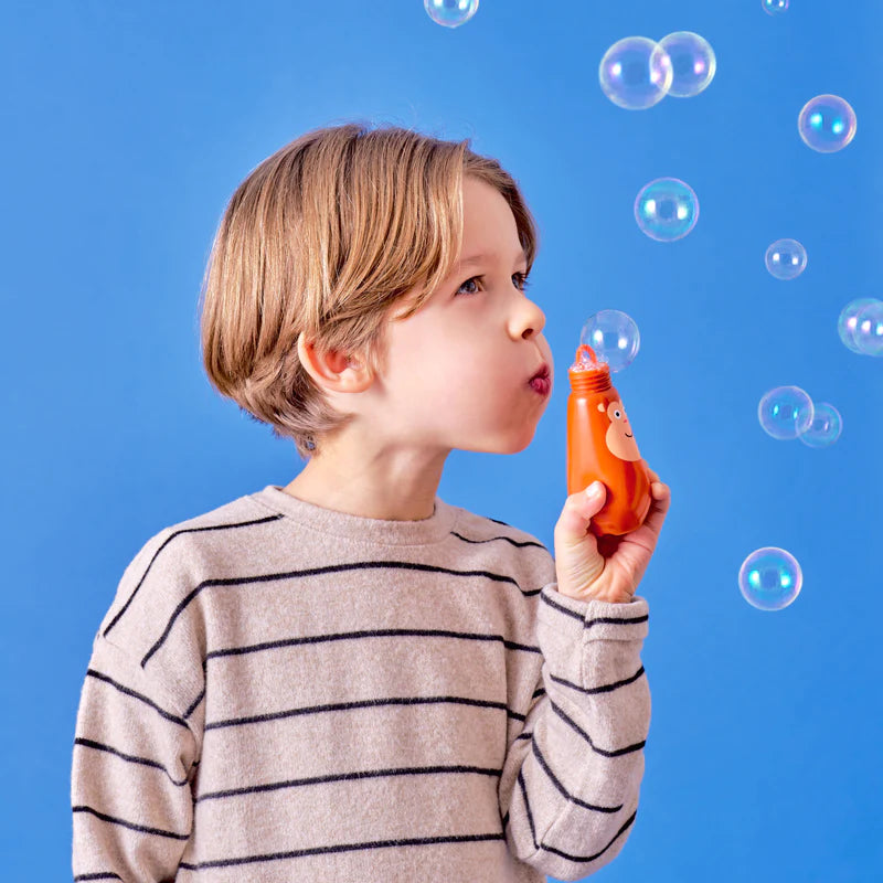 Child blowing bubbles with an orange Squeezy Bubbles toy against a blue background, creating floating bubbles.