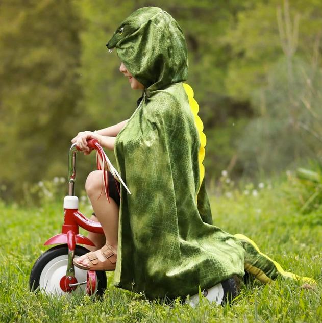 Child wearing the T-Rex Hooded Dinosaur Cape with textured velour and 3D dinosaur details riding a tricycle outdoors.