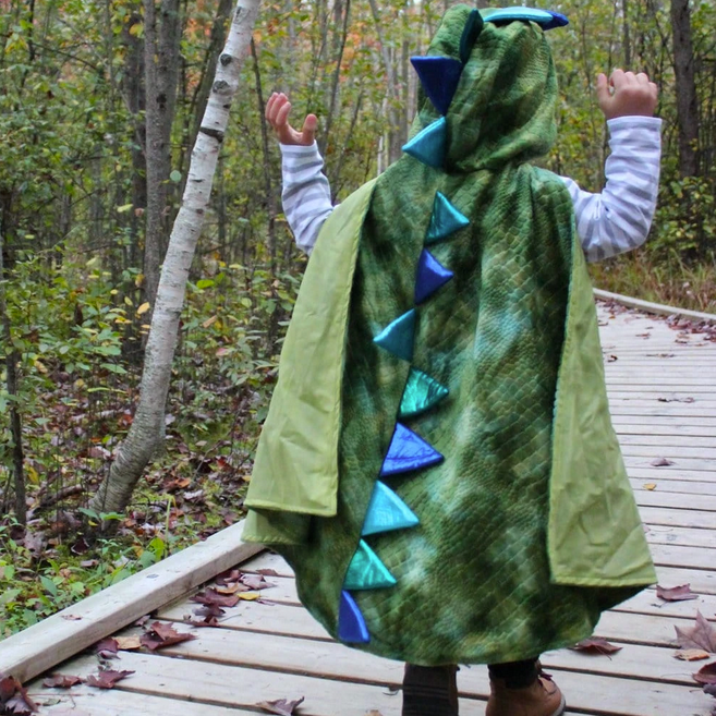 Child wearing a green textured velour Dragon Cape with Claws featuring colorful spikes outdoors on wooden path