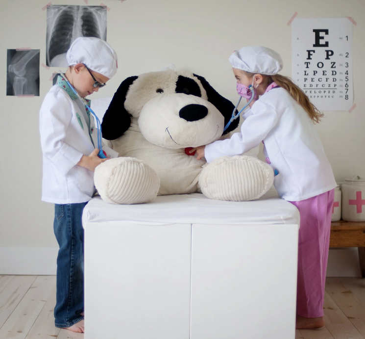 Two kids wearing the Doctor Set costume with white coats and hats play doctor on a large stuffed dog.