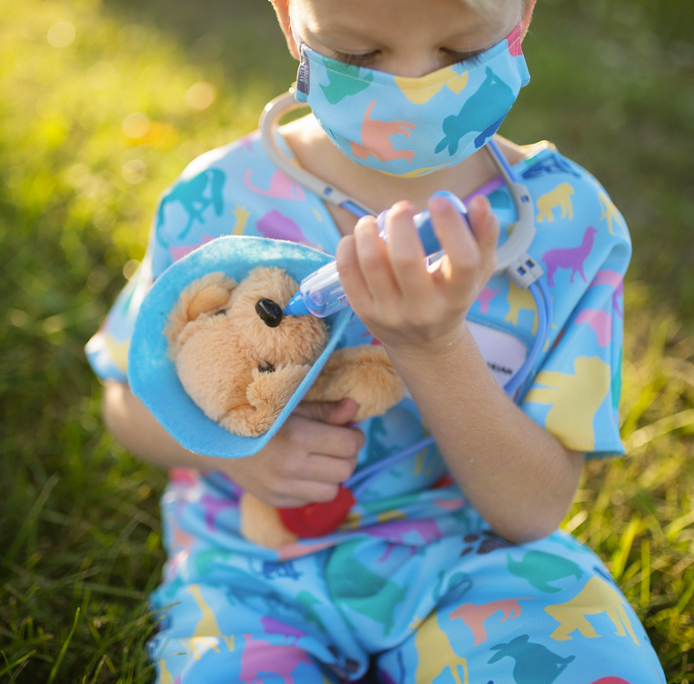 Child dressed in Veterinarian Set playing vet with plush puppy and syringe outdoors in colorful animal print scrubs.