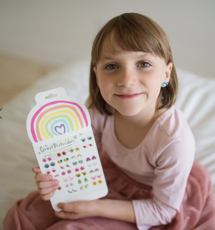 Smiling child wearing blue stick-on earrings holding Stick On Earrings | Rainbow Love pack with colorful designs.