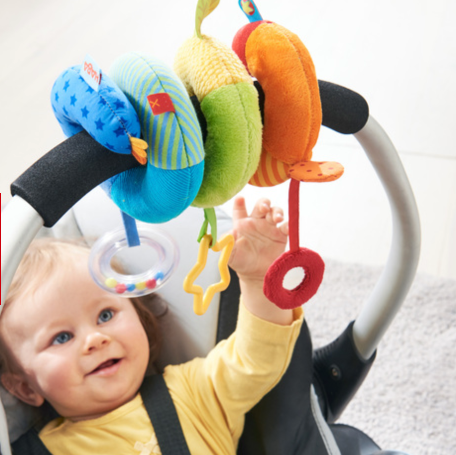 Rainbow Activity Spiral Stroller Toy attached to carrier handle with baby reaching colorful soft rings and shapes.