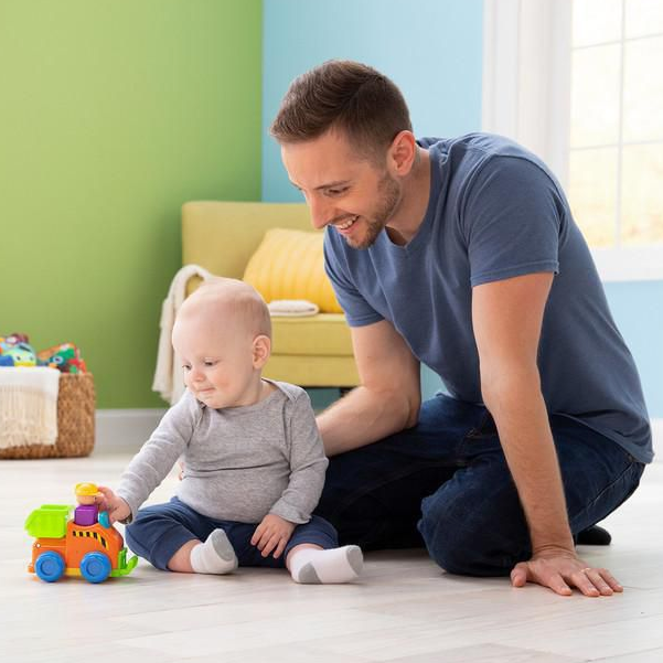 Baby playing with the Press & Go Dump Truck toy while dad watches happily on a bright playroom floor.