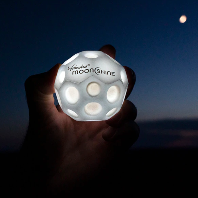 Hand holding glowing Waboba Moon Ball | Moonshine against dark night sky with visible moon in background.