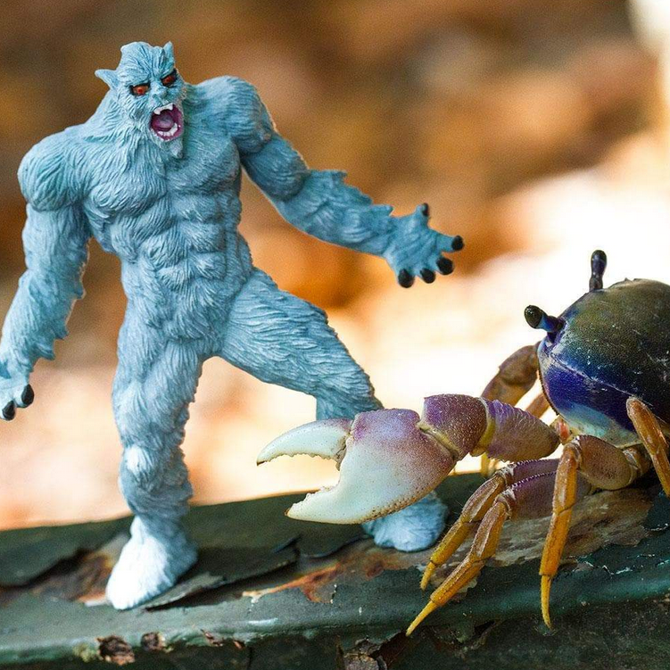 Yeti figure in an intimidation pose with detailed light blue fur facing a crab on a natural surface.