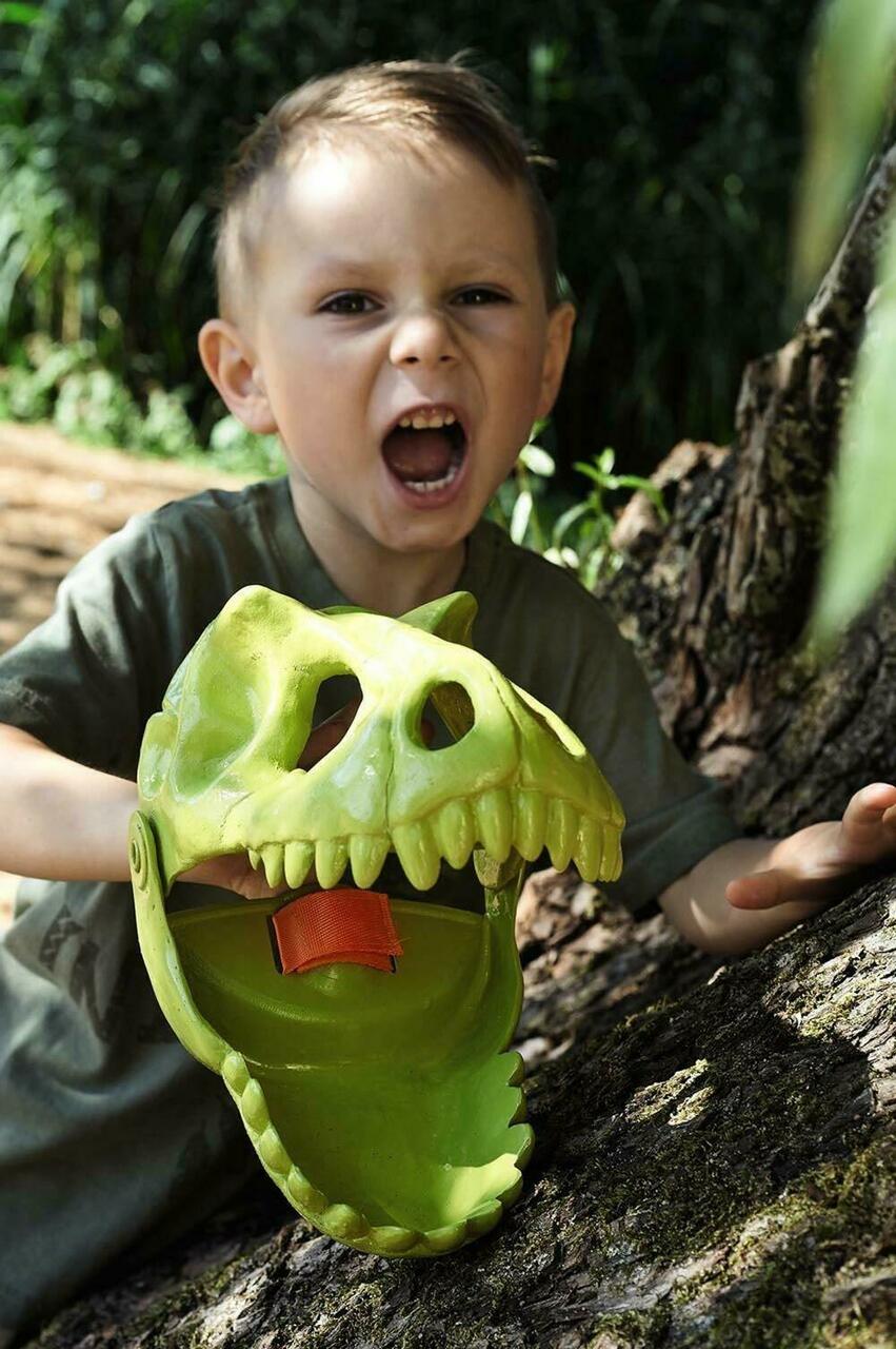 Child playing outdoors holding a green Dinosaur Sand Glove shaped like a dinosaur head for digging and scooping sand.