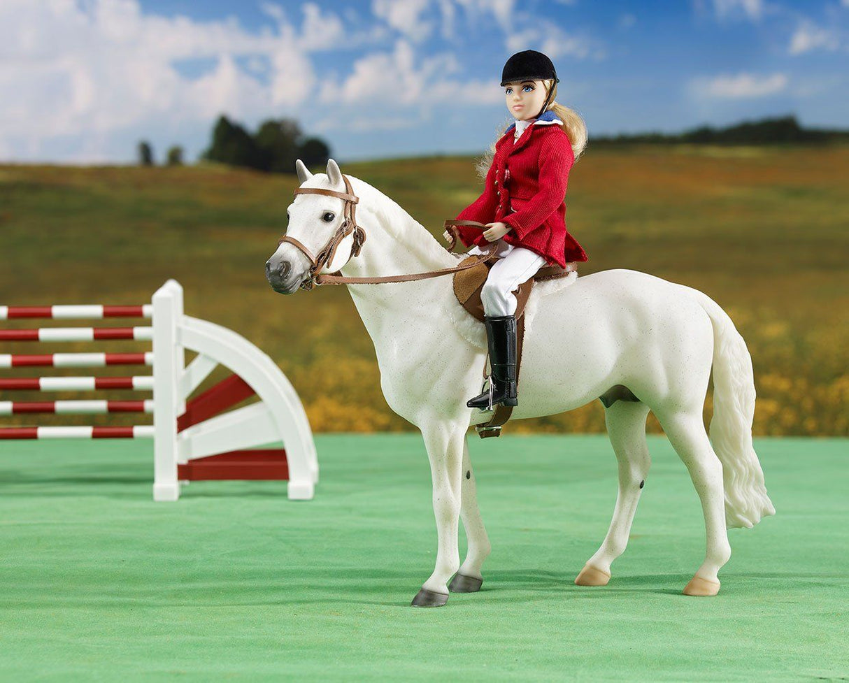 Show Jumper doll in red coat riding white horse over green course with show jumping obstacles in background.
