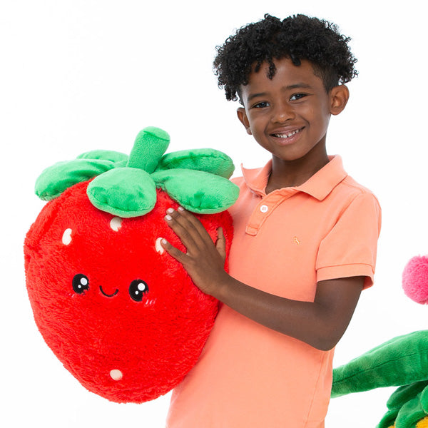 Child smiling and holding a large plush strawberry toy named Strawberry with green leaves and cute face.