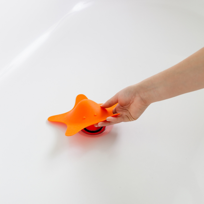 Hand placing the orange STAR Drain Cover over a bathtub drain with a smooth white tub surface visible.