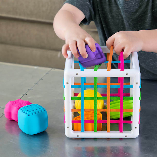 Child playing with Inny Bin shape-sorting cube featuring colorful textured shapes and elastic bands for exploration.