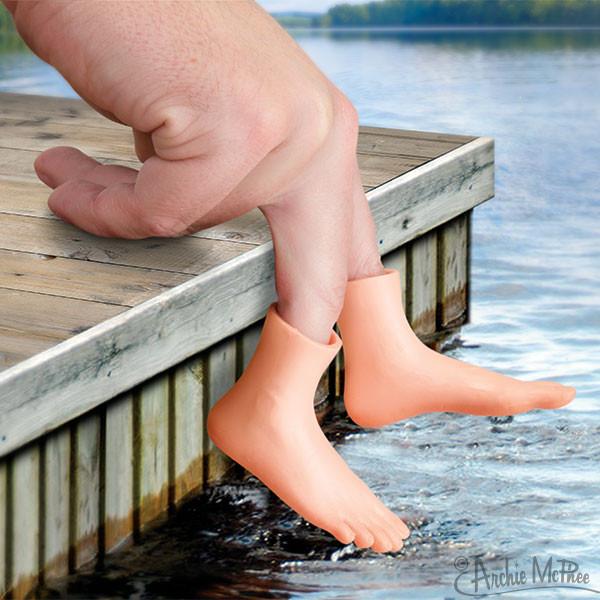 Hand wearing Finger Foot product showing tiny vinyl feet dangling over a wooden dock by water.