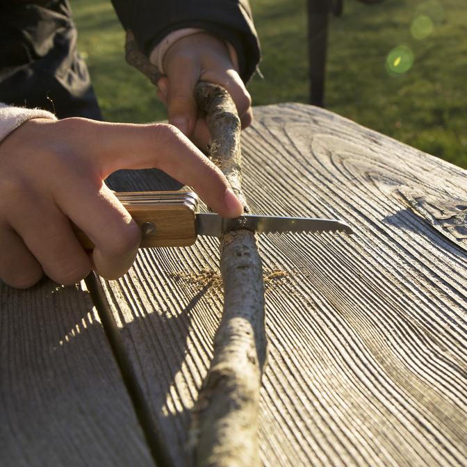 Person using Huckleberry First Pocket Knife saw blade to cut a stick on a wooden surface outdoors.