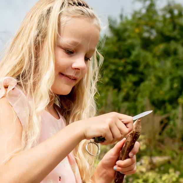 Young girl outdoors using the Huckleberry First Pocket Knife to whittle a wooden stick safely.