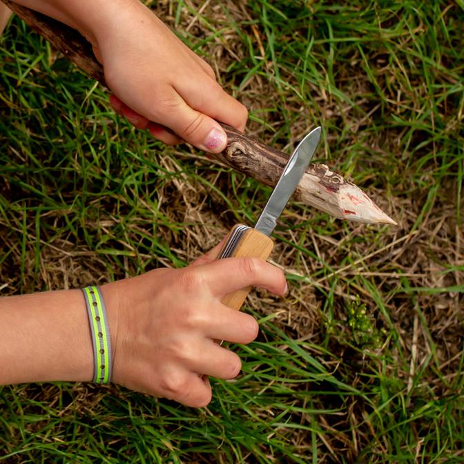 Young hands use the Huckleberry First Pocket Knife to carve a stick outdoors in green grass.