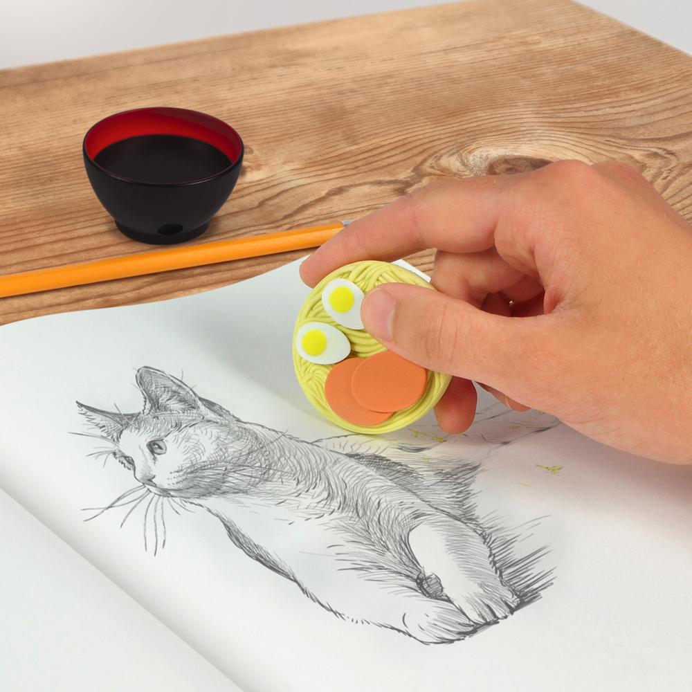 Hand using Ramen Doodles Eraser & Sharpener to erase a pencil sketch of a cat on white paper.