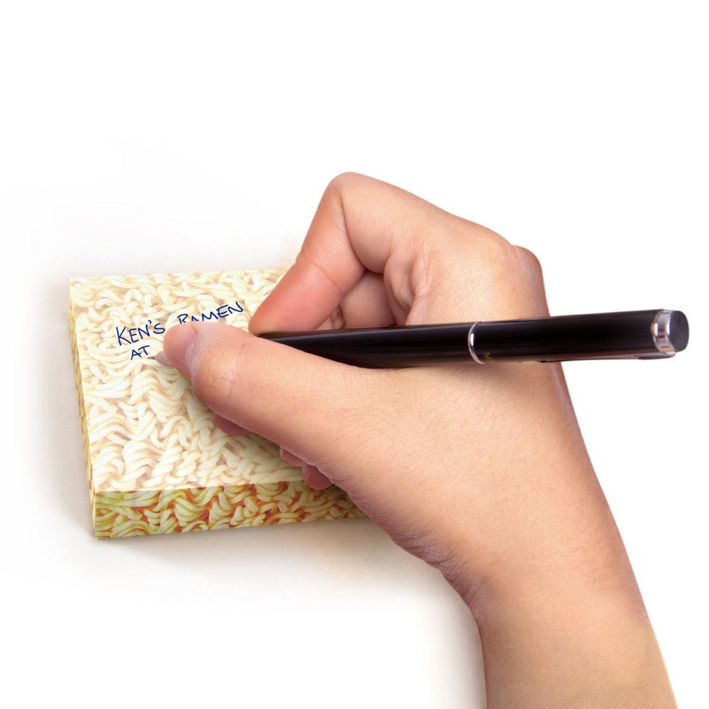 Hand writing a note on Ramen Notes sticky pad designed like ramen noodle packaging for fun memos.