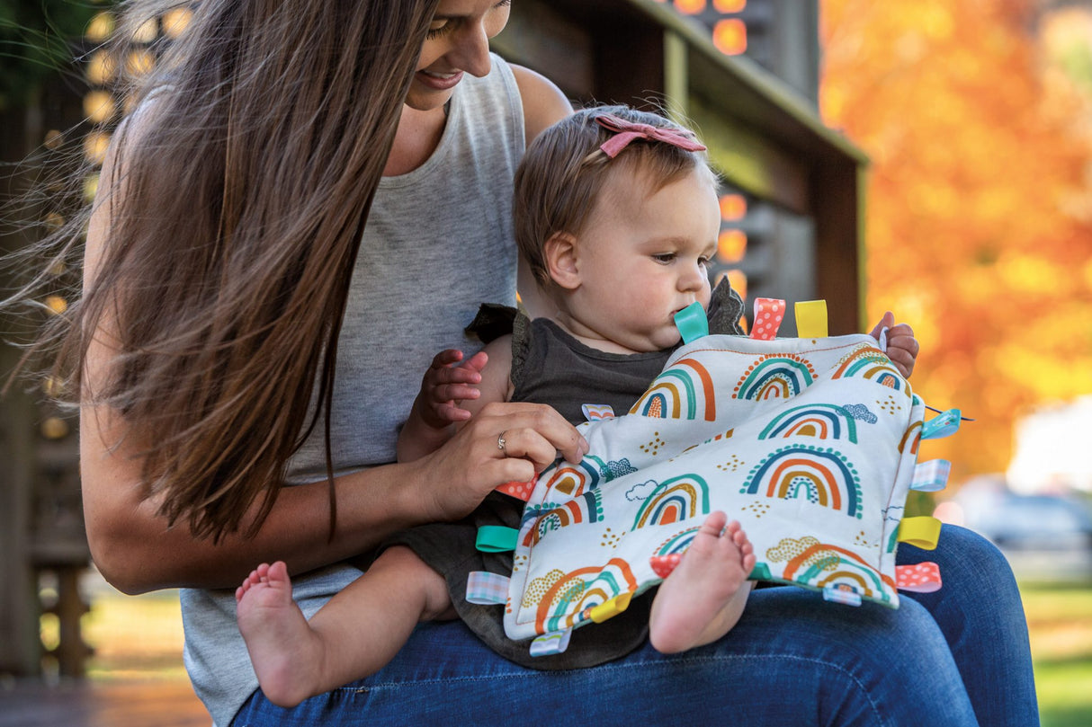Baby playing with Taggies Blankie Rainbow featuring colorful looped ribbons and soft plush backing held by a smiling mom outdoors