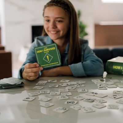 Smiling girl holding a scenario card playing The How I Survived Game surrounded by object chips on a table.