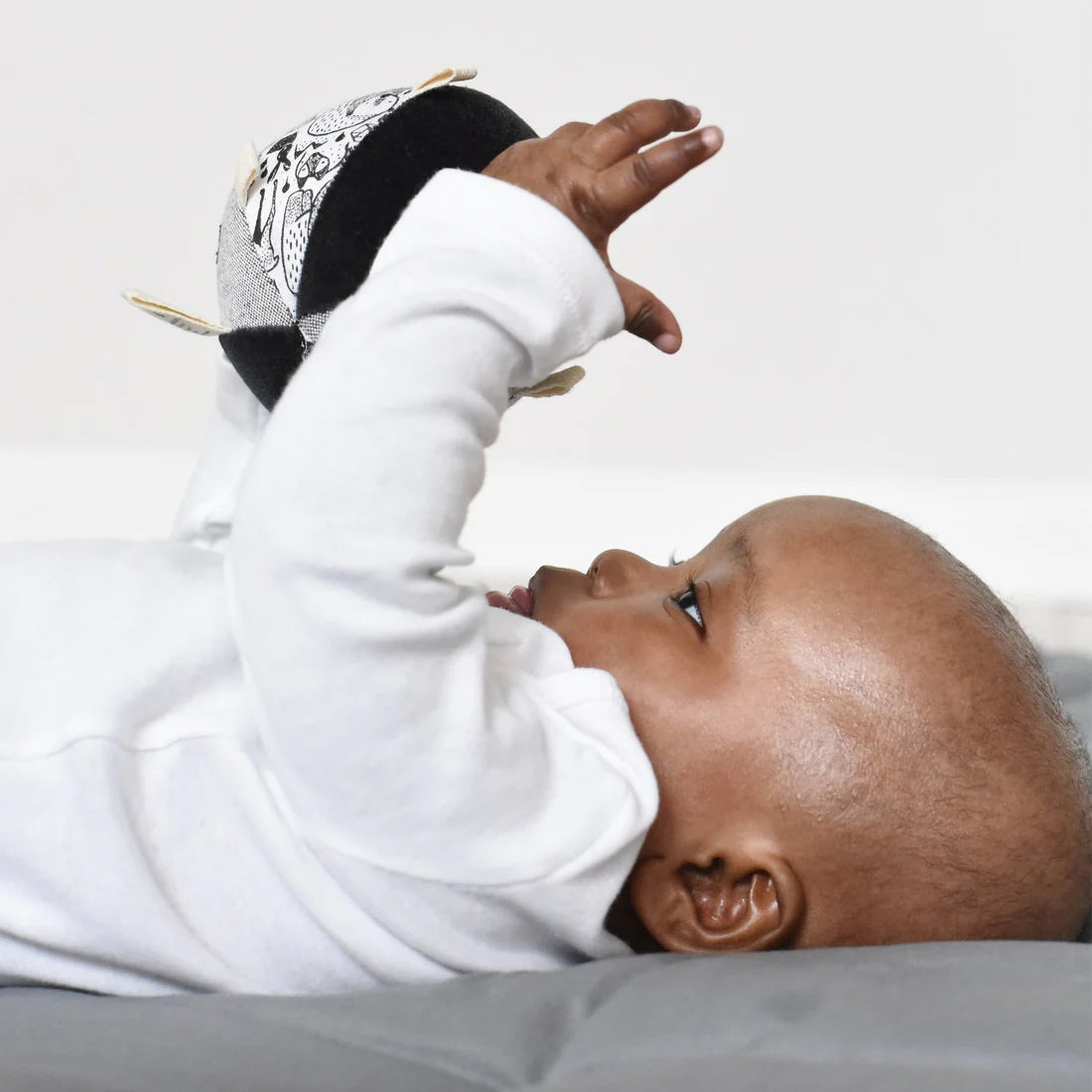 Baby lying on back reaching for Organic Taggy Ball Nordic with high-contrast hand painted graphics and textured fabric.