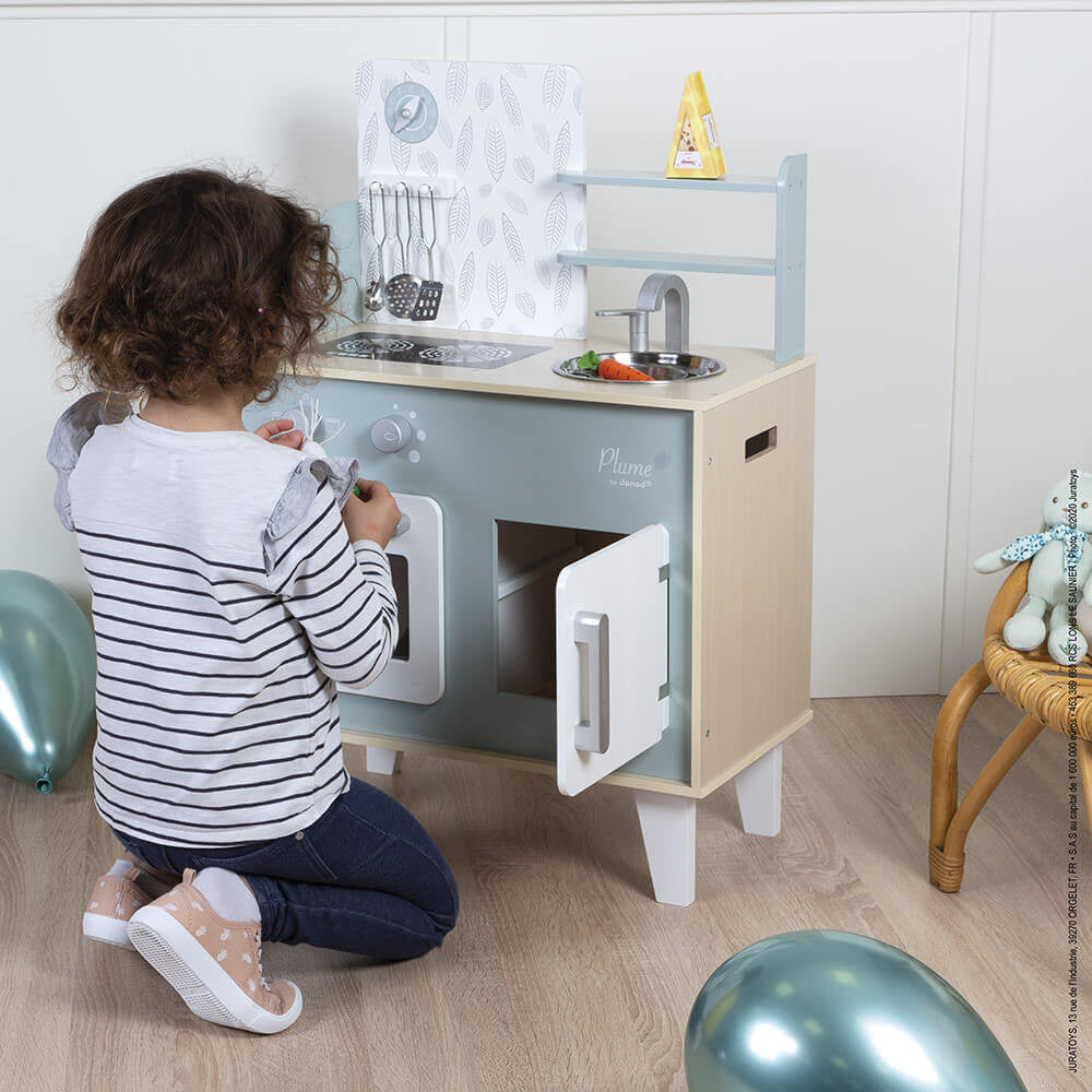 Child playing with Plume Kitchen Cooker wooden toy kitchen set featuring oven, sink, and cooking accessories in blue and grey.