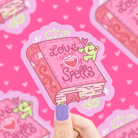 Hand holding a vinyl sticker of a pink love spells book with a smiling green frog and heart designs on a pink background