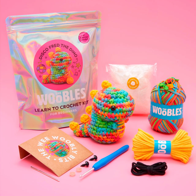 Colorful crochet kit with a rainbow dinosaur, yarn, crochet hook, stuffing, and accessories on a pink background