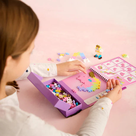 Child assembling a pastel unicorn with Plus Plus Activity Pad | Fairy Tale and storage box on pink surface.
