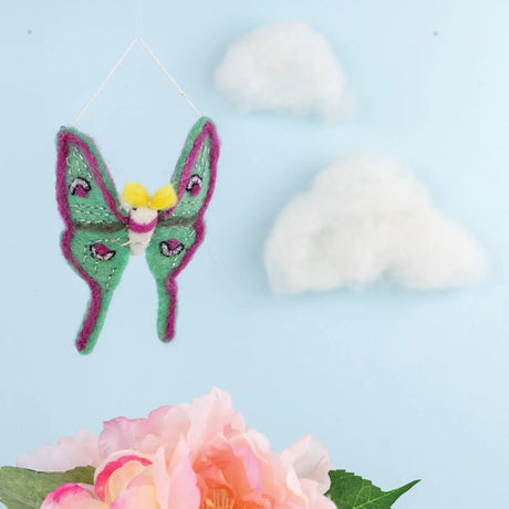 Luna Moth Felt Ornament with green and purple wings, hanging against a blue sky backdrop with clouds and flowers.