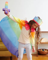 Child wearing a rainbow cape and pastel crown holding a soft rainbow sword while playing indoors