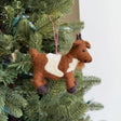 Handmade felt goat ornament in brown and white wool hanging on a Christmas tree branch with red string.