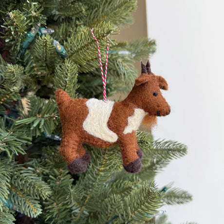 Handmade felt goat ornament in brown and white wool hanging on a Christmas tree branch with red string.