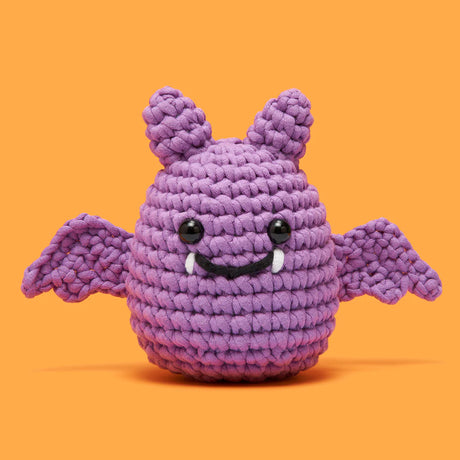 Cute purple crocheted bat with wings, black eyes, and small white fangs on an orange background