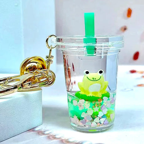 Clear plastic cup with green straw featuring a cute frog and glitter inside, Frog Boba Floaty Keychain attached to gold keyring