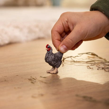 Hand holding a small, detailed Plymouth Rock Chicken figurine with black and white striped feathers on a wooden surface.