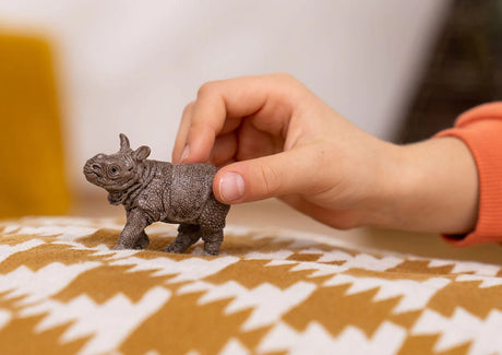 Child’s hand holding a detailed Indian Rhinoceros Baby figurine with thick scaly skin on patterned fabric surface.
