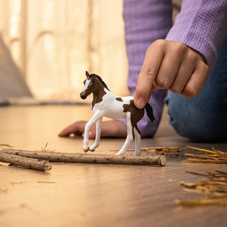 Paint Horse Foal figurine with brown and white coat pattern, black mane, and tail held by a child's hand on the floor.