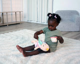 Toddler girl in green plays on white rug feeding soft doll with pastel pink and yellow cup and spoon