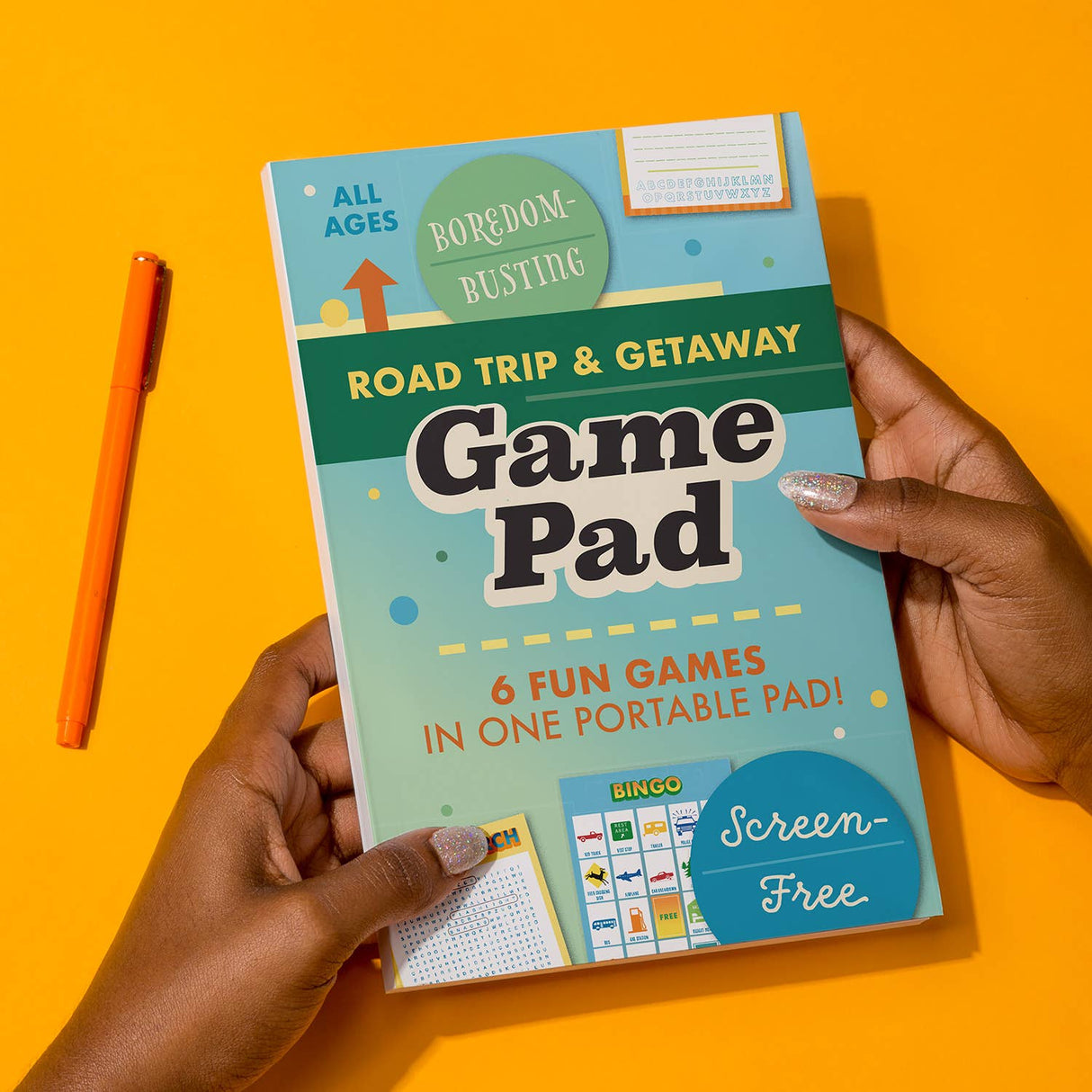 Hands holding a colorful Road Trip and Getaway Game Pad with orange pen on yellow background