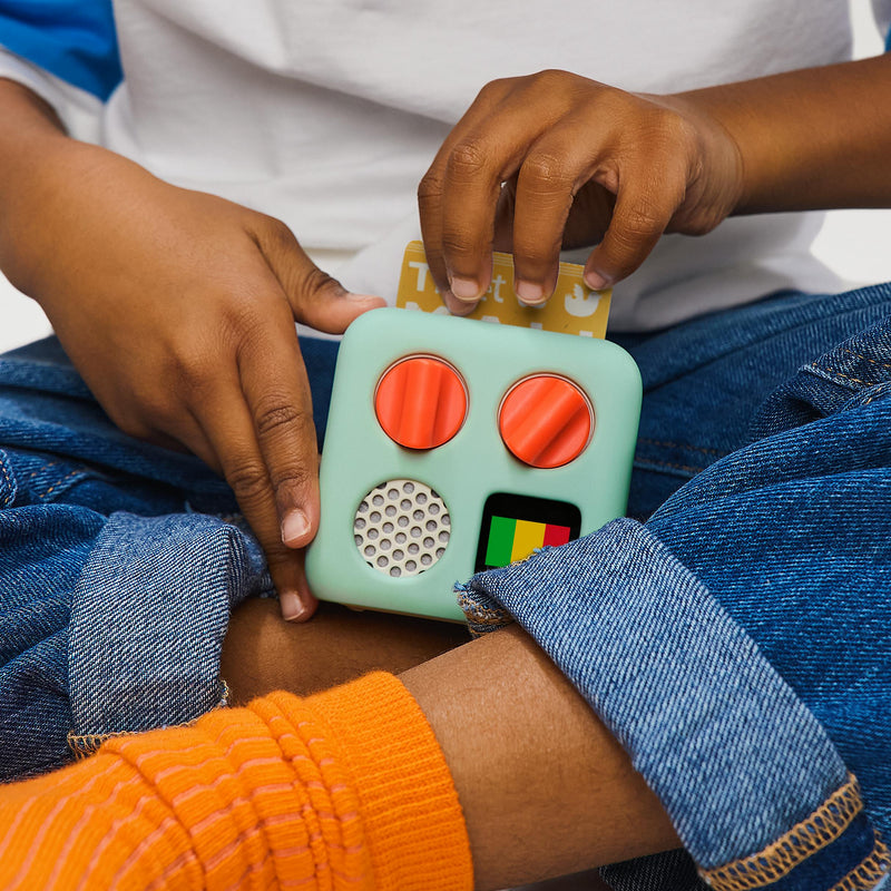 Child holding a small mint green music player with orange dials while sitting cross-legged wearing denim and orange socks