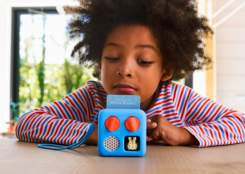 Child watching a small blue audio device playing a story with a card inserted on a bright day indoors