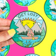 Get Lost Wilderness Badge Sticker showing a cartoon bear paddling a canoe with mountains and sun background.
