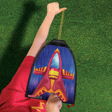 Child holding a vibrant rubber band rocket kite with red and yellow rocket design ready to fly over green grass.