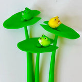 Three colorful frog gel pens resting on bright green lotus leaf tops against a plain background