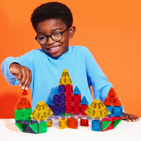 Child playing with colorful Magnatiles Micromags magnetic building blocks on white table against orange background