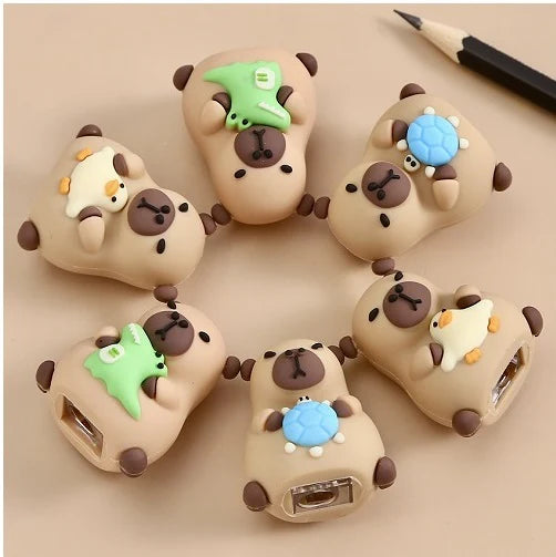 Six cute capybara-shaped pencil sharpeners with small animal decorations on a beige background next to a pencil.