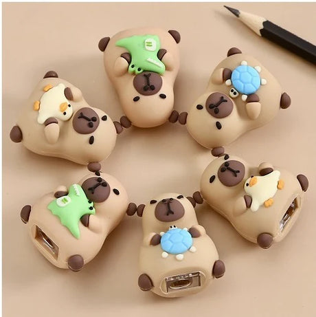 Six cute capybara-shaped pencil sharpeners with small animal decorations on a beige background next to a pencil.